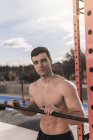 Junger Mann ohne Hemd lehnt an der Bar und schaut in die Kamera, während er an einem sonnigen Tag auf dem Sportplatz in der Stadtstraße trainiert — Stockfoto