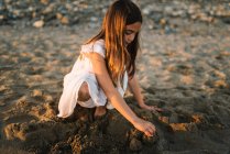Cute pensive female kid in white dress playing with sand on seaside in sunlight — Stock Photo