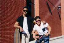 Joven feliz afroamericano guapo hombres en ropa casual y gafas de sol de pie en la calle con bicicleta y monopatín - foto de stock