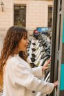 Smiling young lady in casual outfit using kiosk on bicycle sharing station on city street — Stock Photo