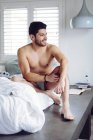 Jovem sorrindo bem sucedido sexy homem com penteado elegante em cuecas cinza deitado na cama em casa e olhando para longe — Fotografia de Stock