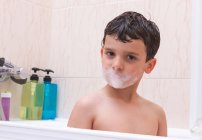Очаровательный ребенок смотрит в камеру с покрытым пеной ртом, сидя в ванной комнате — стоковое фото