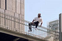 Junger afrikanisch-amerikanischer Mann sitzt an einem sonnigen Tag auf dem Geländer einer Brücke an der Stadtstraße — Stockfoto