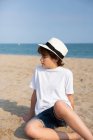 Cute boy in white t-shirt and hat sitting on sandy beach in summer day — Stock Photo