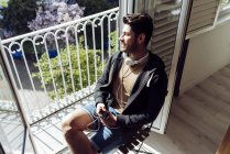 Young smiling handsome casual male in headset leaning on terrace railing with smartphone and looking away — Stock Photo