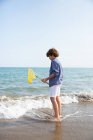 Side view of long hair cute boy in summer wear standing in water with fishing net on sea shore in sunny day — Stock Photo