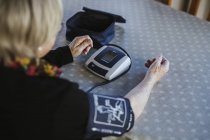 Mujer mayor usando tensiómetro para medir la presión arterial mientras está sentada en la mesa en casa - foto de stock
