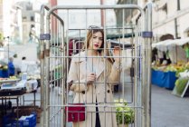 Молодая чувственная женщина в стильном наряде смотрит на камеру через решетку на рынке на улице — стоковое фото