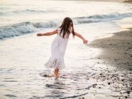 Маленька дівчинка в білій сукні грає на березі моря на заході сонця — стокове фото