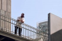 Jeune homme afro-américain appuyé sur la rampe du pont sur la rue de la ville par une journée ensoleillée — Photo de stock