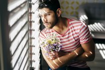 Jovem estressado sozinho macho em roupas casuais sentado na cadeira com buquê de flores e olhando para fora janela — Fotografia de Stock