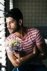 Joven estresado solo hombre en ropa casual sentado en la silla con ramo de flores y mirando por la ventana - foto de stock