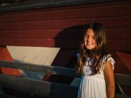Портрет милой маленькой девочки в белом платье, опирающейся на деревянную стену при солнечном свете — стоковое фото