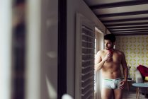 Side view of young pensive sexy handsome male in underwear standing near terrace at home and looking away — Stock Photo