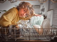 Хлопчик посміхається, дивлячись всередину відкритої посудомийної машини на кухні — стокове фото