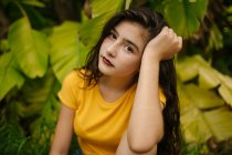 Задумчивая молодая женщина в желтой футболке поддерживает голову и смотрит в камеру, сидя рядом с экзотическим кустарником в саду — стоковое фото