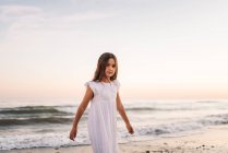 Petite fille en robe blanche marchant et regardant la caméra sur le bord de la mer au coucher du soleil — Photo de stock