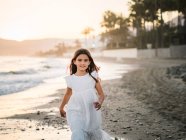 Весёлая милая девочка в белом платье ходит по песчаному побережью и смотрит в камеру — стоковое фото