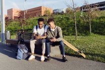 Jovens afro-americanos felizes amigos masculinos bem sucedidos em roupas casuais e tênis sentados no banco e navegando smartphone juntos — Fotografia de Stock