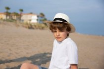 Lindo chico en camiseta blanca y sombrero sentado en la playa de arena en el día de verano - foto de stock