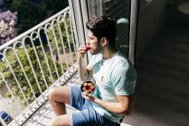 Vista lateral de un joven alegre y sexy hombre exitoso en ropa interior sentado en una silla de madera y comiendo fresa cerca del balcón - foto de stock
