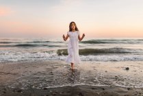 Menina em vestido branco andando na água na praia ao pôr do sol — Fotografia de Stock
