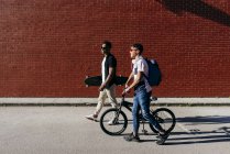 Vista lateral de jóvenes negros alegres amigos varones exitosos en gafas de sol caminando en la ciudad con bicicleta y monopatín - foto de stock