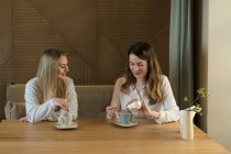 Deux copines élégantes avec des tasses de café frais souriant et parlant tout en étant assis à la table dans un restaurant confortable — Photo de stock
