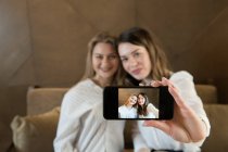 Deux copines élégantes avec des tasses de café frais souriant et posant pour selfie tout en étant assis à la table dans un restaurant confortable — Photo de stock