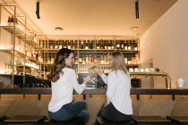 Вид ззаду на двох друзів-жінок, посміхаючись і кмітливі келихи алкогольних коктейлів, проводячи час у барі затишного ресторану — стокове фото
