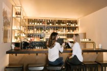 Обзор двух подруг, улыбающихся и звенящих стаканов алкогольных коктейлей во время пребывания в баре уютного ресторана — стоковое фото