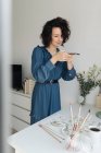 Жінка в блакитній сукні знімає на мобільному телефоні акварельні роботи на столі в студії — стокове фото