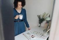 Brunette artiste féminine debout à côté d'une table avec une tasse de café et regardant le dessin dans le lieu de travail — Photo de stock