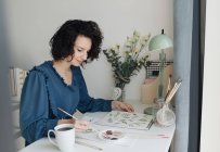 Femme élégante avec pinceau peinture aquarelle fleurs sur feuille au bureau — Photo de stock