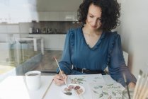 Blick durch ein Fenster einer eleganten Frau mit Pinsel, die Aquarellblumen auf Laken am Schreibtisch malt — Stockfoto