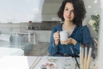 Красивая брюнетка-художница сидит за столом с чашкой кофе и смотрит в сторону на рабочем месте — стоковое фото