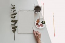 Vista superior da mão pessoa com pincel pintura aquarela plantas na folha em branco na mesa — Fotografia de Stock