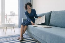 Beautiful young brunette female sitting on sofa and looking album with drawings at home — Stock Photo