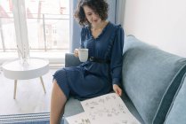 Beautiful young brunette female sitting on sofa and looking album with drawings at home while enjoying a cup of coffee — Stock Photo
