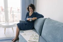 Beautiful young brunette female sitting on sofa and looking album with drawings at home while enjoying a cup of coffee — Stock Photo
