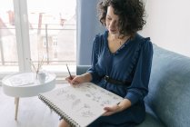 Elegante Frau, die zu Hause auf einem Sofa sitzt und auf einem Notizbuch zeichnet — Stockfoto
