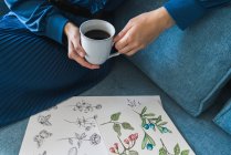 Cropped view of unrecognizable female artist sitting on sofa and looking album with drawings at home while enjoying a cup of coffee — Stock Photo