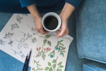 Обрезанный вид неузнаваемой художницы, сидящей на диване и смотрящей альбом с рисунками дома, наслаждаясь чашкой кофе — стоковое фото