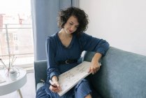 Elegante Frau, die zu Hause auf einem Sofa sitzt und Blumen auf einem Notizbuch zeichnet — Stockfoto
