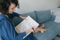 Elegante Frau, die zu Hause auf einem Sofa sitzt und Blumen auf einem Notizbuch zeichnet — Stockfoto