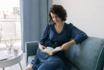 Щаслива молода брюнетка в блакитній сукні читає книгу, сидячи на дивані за столом з творчими аксесуарами та талісманами та альбомом вдома — стокове фото