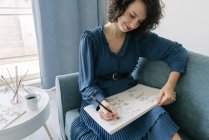 Elegant woman sitting on a sofa drawing flowers on a notebook at home — Stock Photo