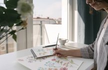 Abgeschnitten von Person Hand mit Pinsel Malerei Aquarell Blumen auf großem Blatt am Schreibtisch — Stockfoto