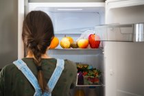 Невизначена жінка бере свіжі фрукти з полиці холодильника вдома — стокове фото