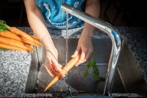 Abgeschnittenes Bild einer Frau, die zu Hause reife Karotten unter sauberem Wasser über der Spüle wäscht — Stockfoto